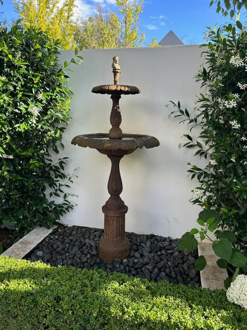Ornamental bird bath fountain on a wooden pedestal among green plants and white flowers